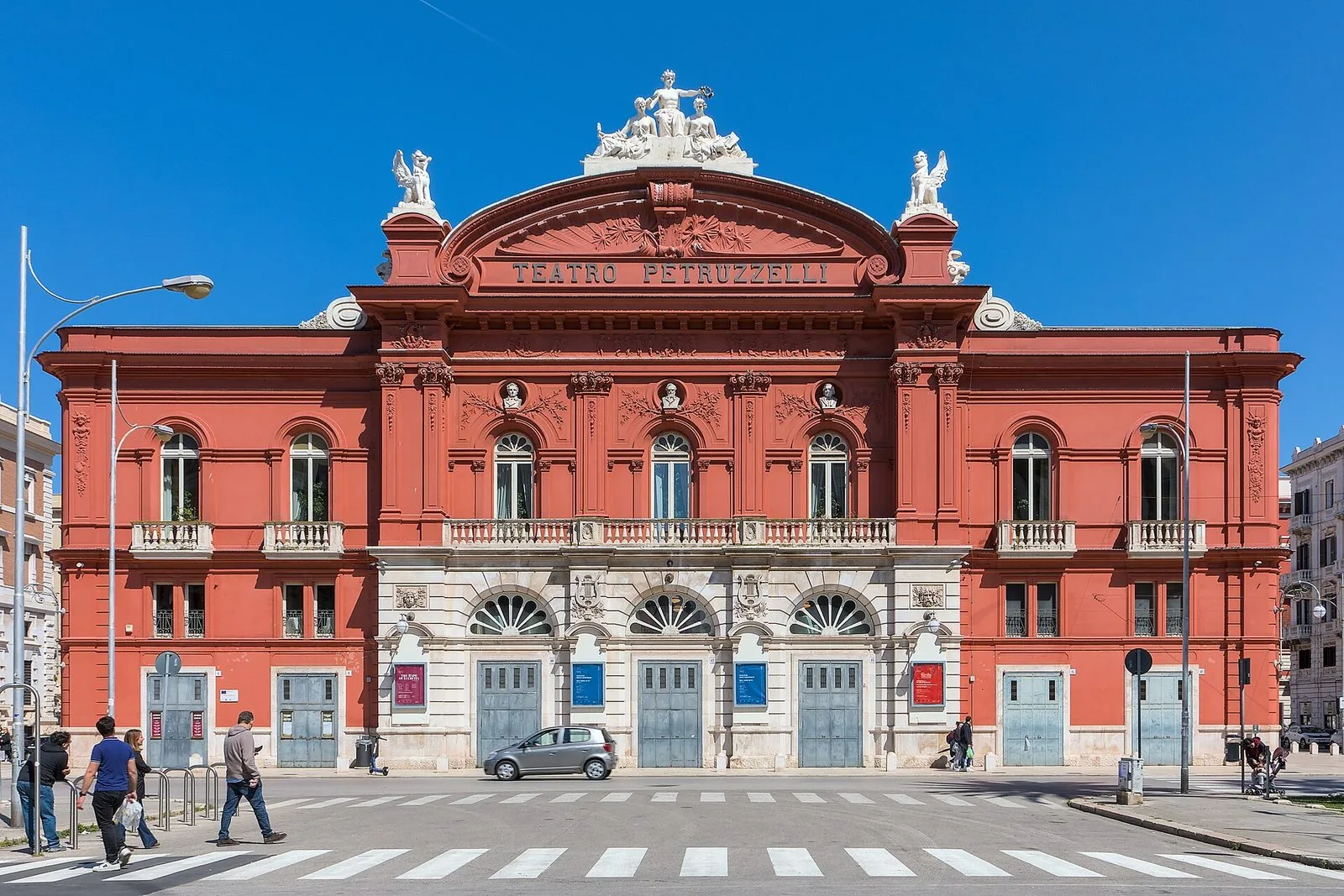 La facciata rossa del Teatro Petruzzelli di Bari con le statue sul coronamento e la scritta del teatro, sotto un cielo azzurro