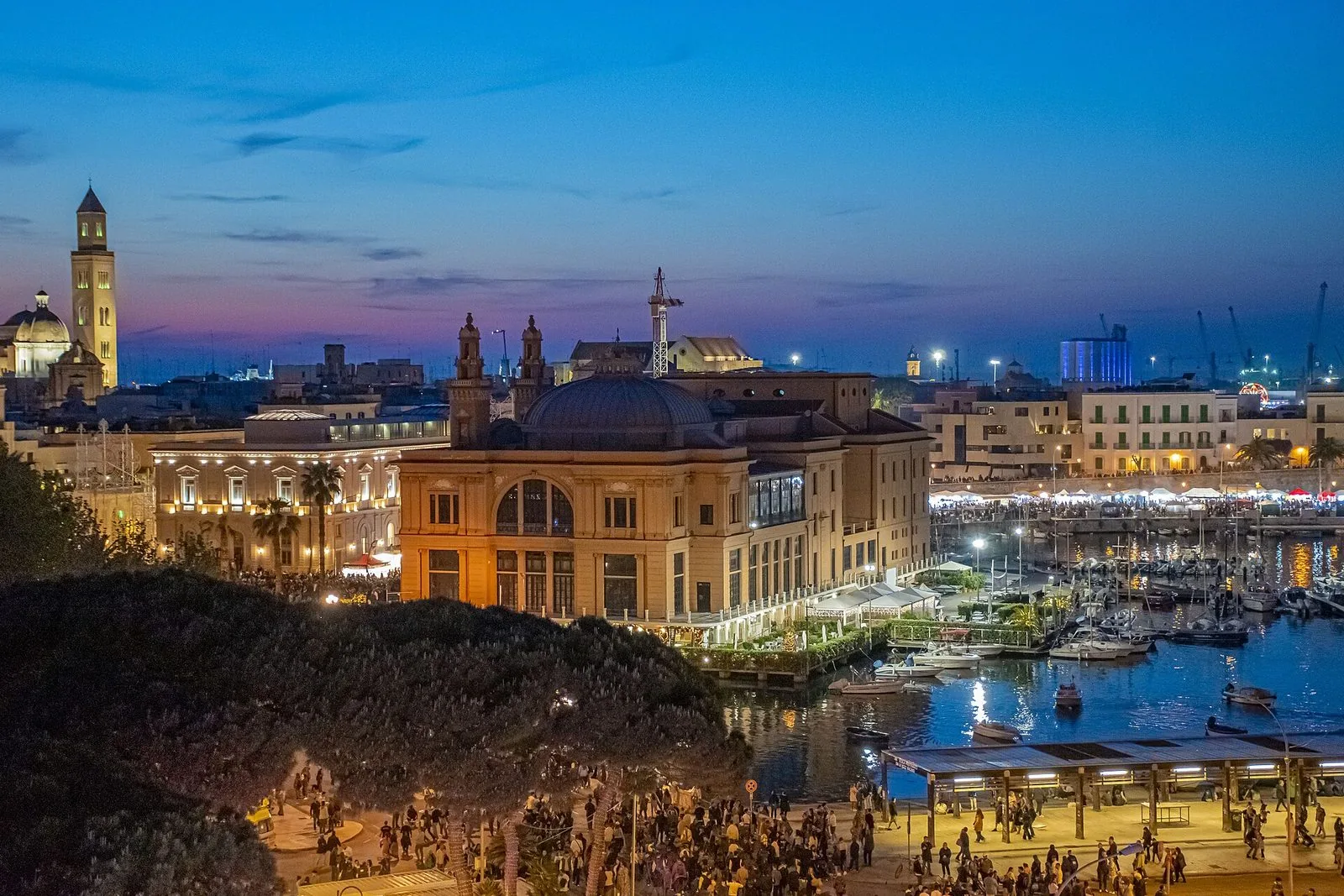 Bari al crepuscolo durante la festa di San Nicola: il Teatro Margherita sul porticciolo, i campanili della città vecchia e la folla sul lungomare